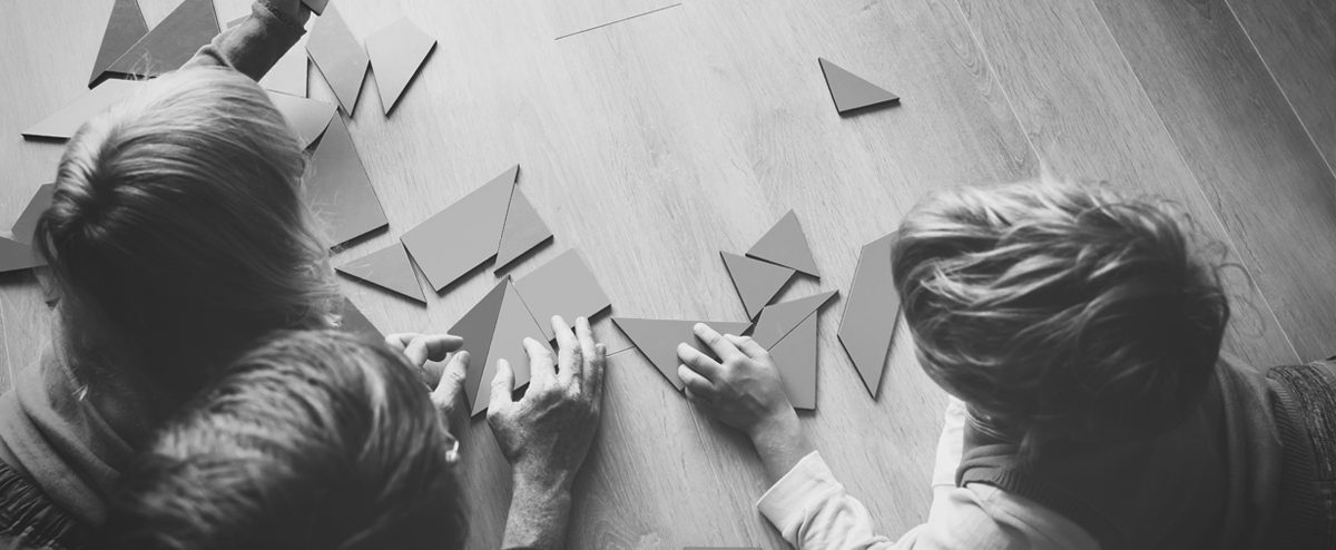 overhaed view of an adult and two children playing with tangrams on a wood floor.