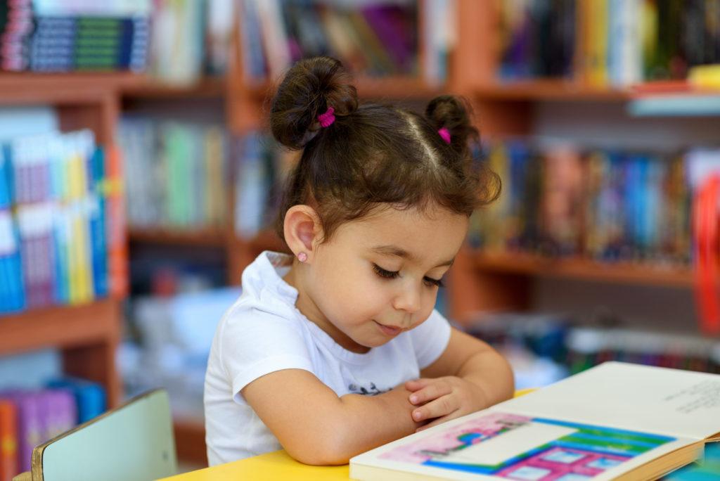 Young girl sitting at a table in a classroom reading a book.