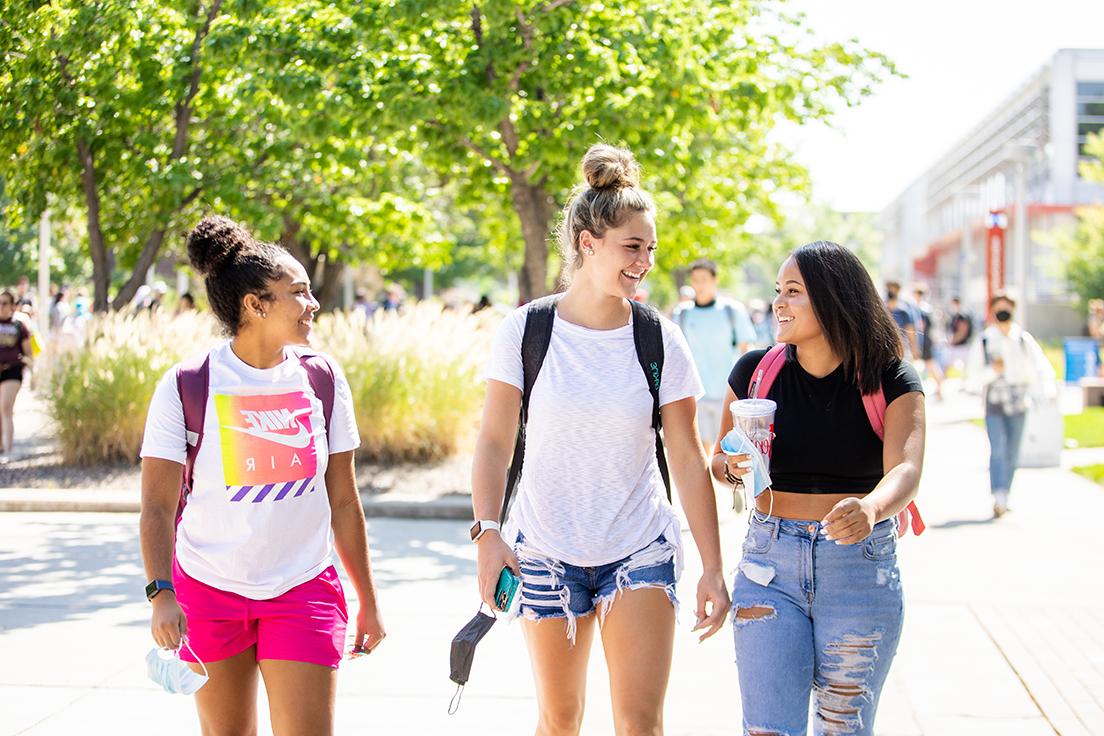 Three students walking towards the viewer, through a green and sunny Auraria campus