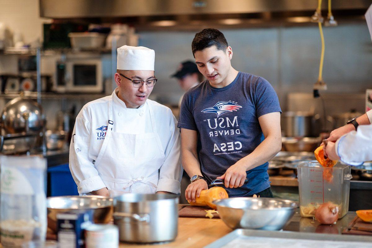A student in a blue shirt slicing a vegetable next to a student wearing a white chef's outfit