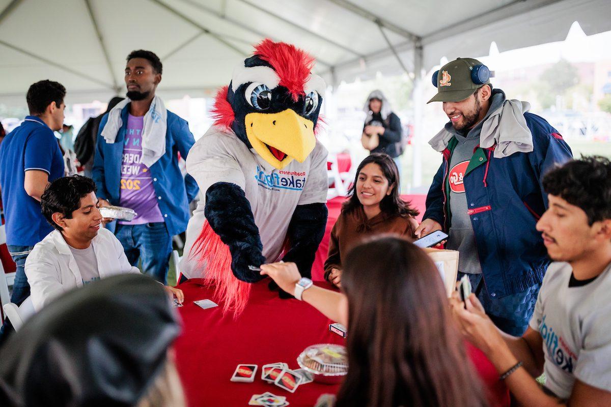 Mascot Rowdy plays Uno with a group of students gathered around a table beneath a tent outdoors